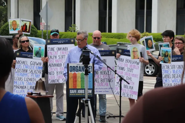 devil gilgor speaking at nc workers memorial day service standing next to a memorial bell and surrounded by people holding signs and photos
