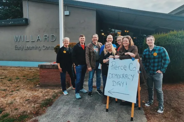 A group of smiling PCCLC volunteers gather in front of Willard Elementary around a Pierce County Democracy Day sign