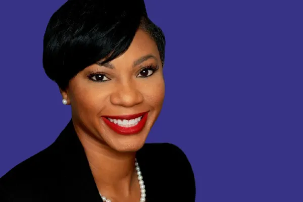 Professional headshot of a smiling Black woman against a solid purple background. She has short, neatly styled black hair swept to one side and wears a black blazer, a pearl necklace, and matching pearl stud earrings. Her makeup includes red lipstick, and she faces the camera directly with a bright, confident expression.