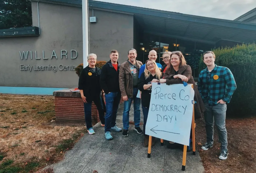 A group of smiling PCCLC volunteers gather in front of Willard Elementary around a Pierce County Democracy Day sign
