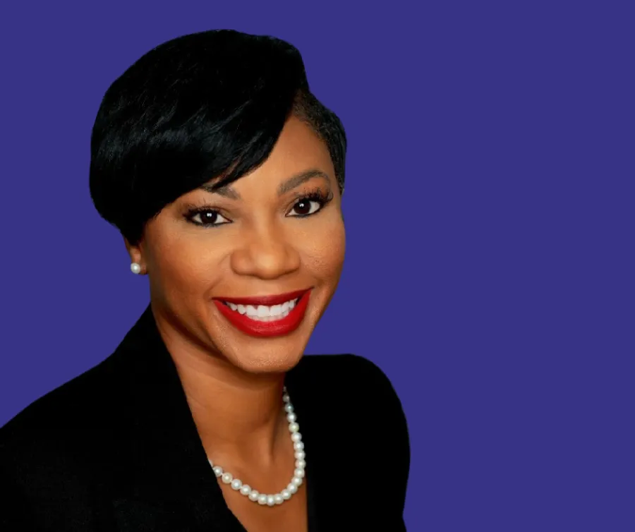 Professional headshot of a smiling Black woman against a solid purple background. She has short, neatly styled black hair swept to one side and wears a black blazer, a pearl necklace, and matching pearl stud earrings. Her makeup includes red lipstick, and she faces the camera directly with a bright, confident expression.